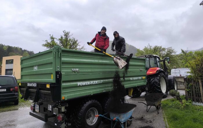 Zwei Helfer schaufeln bei Regenwetter Komposterde von einem Traktoranhänger in eine Schubkarre.